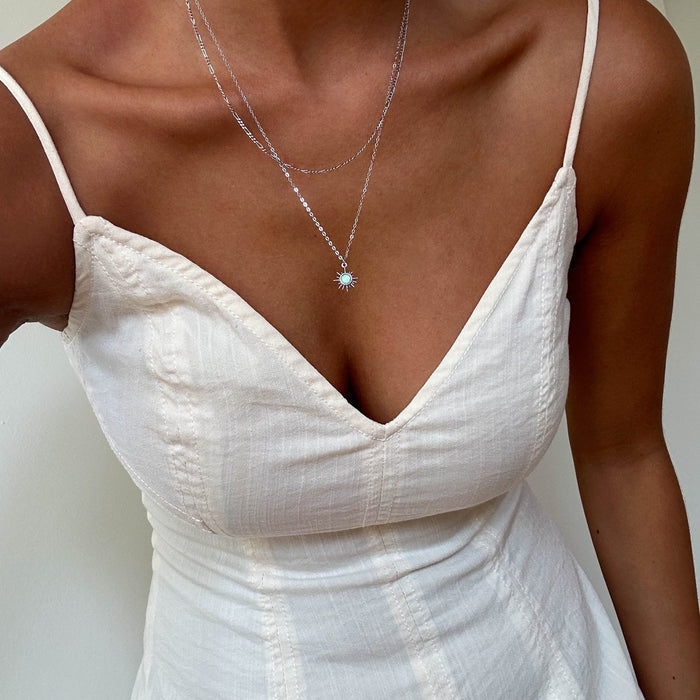A person wearing a cream, sleeveless dress with thin straps and a deep V neckline, shown from the shoulders down. They accessorize with LLUME Jewelry’s Cyra Daily Set Silver and another necklace featuring a sun-shaped pendant.