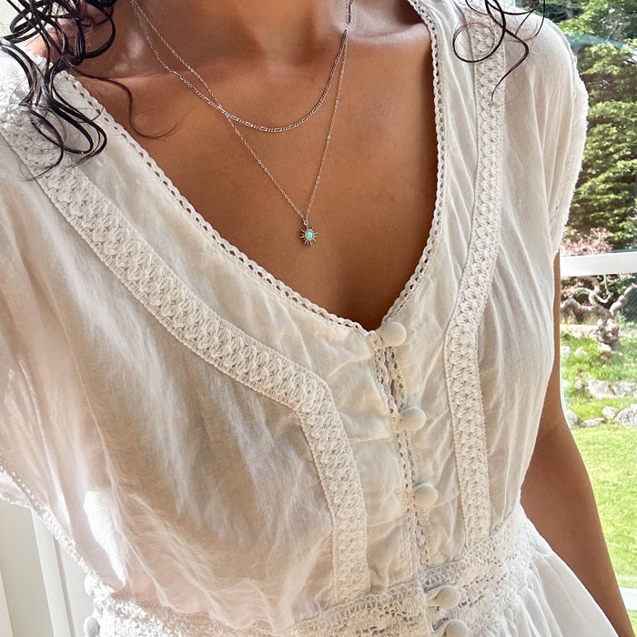 A person stands near a window, sunlight highlighting their white textured dress and layered silver necklaces from LLUME Jewelry, including the Cyra Daily Set Silver; greenery is visible outside.