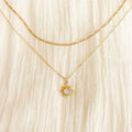 The Cyra Daily Set by LLUME Jewelry features two delicate gold necklaces on cream fabric: a simple chain and the Cyra opal pendant with a sun-shaped charm and opalescent stone—an elegant pairing for effortless style.