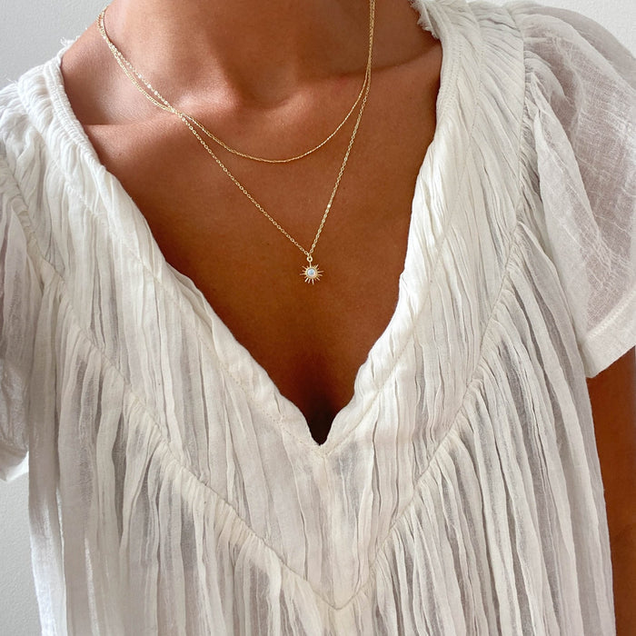 Someone wears a white, pleated V-neck blouse and the LLUME Jewelry Cyra Daily Set—one necklace with a sparkling Cyra opal pendant and another featuring a delicate sunburst design with a central stone.