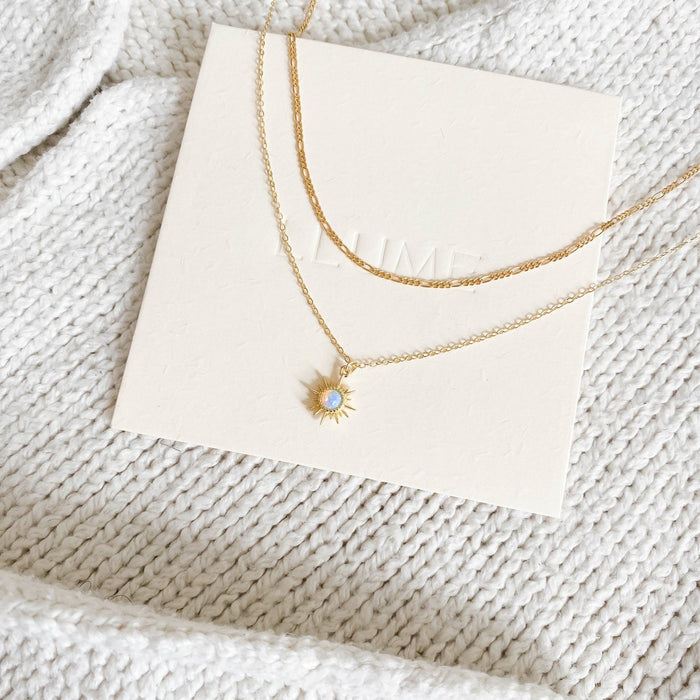 The LLUME Jewelry Cyra Daily Set displays two delicate gold necklaces on a cream card atop textured white knit—a sun-shaped pendant with a light blue stone and a classic Colette necklace.
