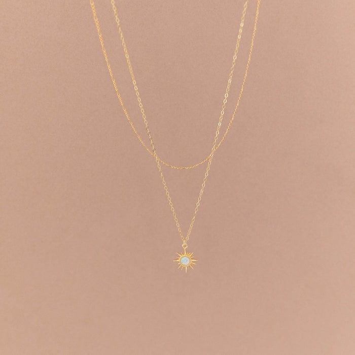 The LLUME Jewelry Cyra Daily Set features a delicate gold necklace with a sunburst pendant and small white stone, displayed on a plain beige background.