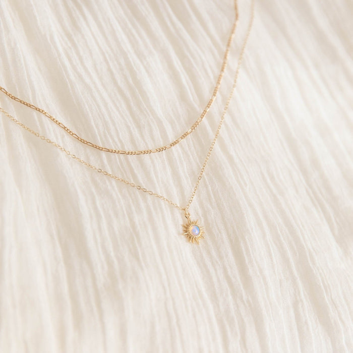 Two delicate gold necklaces from LLUME Jewelry—the Cyra Daily Set—rest on white textured fabric. One features a sun-shaped pendant with a light blue stone, the other is a fine chain. This elegant set adds charm to any look.