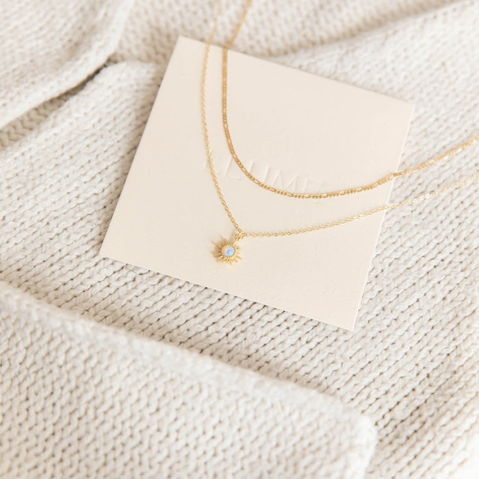 The Cyra Daily Set by LLUME Jewelry features a delicate sun-shaped pendant with a small gemstone, displayed on a cream card and set against a beige knitted fabric background.