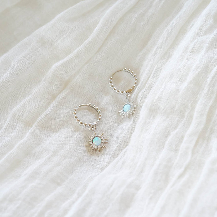 Cyra Hoops Silver by LLUME Jewelry: 925 silver hoop earrings with sunburst pendants and light blue stones, displayed on a softly textured white fabric background.