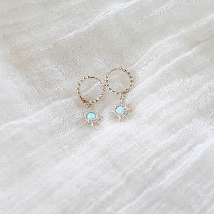 The Cyra Hoops Silver by LLUME Jewelry feature silver beaded hoops with sunburst opal sun charm pendants and iridescent centers, elegantly displayed on textured white fabric.
