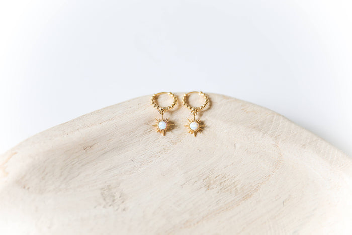 The Cyra Hoops by LLUME Jewelry are gold-plated sterling silver earrings with sun-shaped pendants and white center stones, shown on a light wooden dish against a white background.