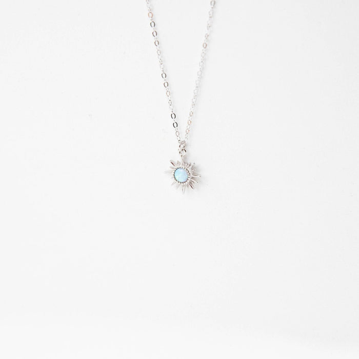 The Cyra Necklace Silver by LLUME Jewelry features a delicate 925 sterling silver chain and a small sun-shaped pendant with a light blue gemstone, shown on a plain white background.