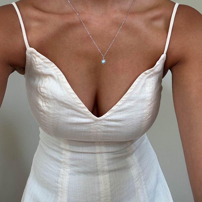 A person in a white, sleeveless dress with thin straps and a deep V-neckline pairs it with LLUME Jewelry’s Cyra Necklace Silver—a layered 925 sterling silver chain featuring a tiny squared white opal charm. The background is plain and neutral.