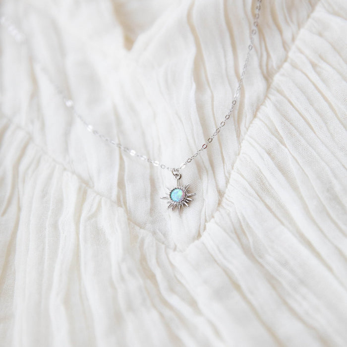 The Cyra Necklace Silver by LLUME Jewelry features a delicate 925 sterling silver chain with a sun-shaped pendant and a tiny square white opal charm, shown resting on softly pleated off-white fabric.