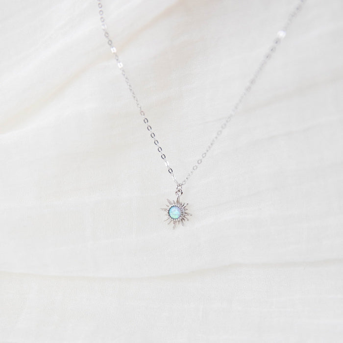 The Cyra Necklace Silver by LLUME Jewelry features a delicate 925 sterling silver chain with a sun-shaped pendant and opalescent center, beautifully displayed against white fabric.