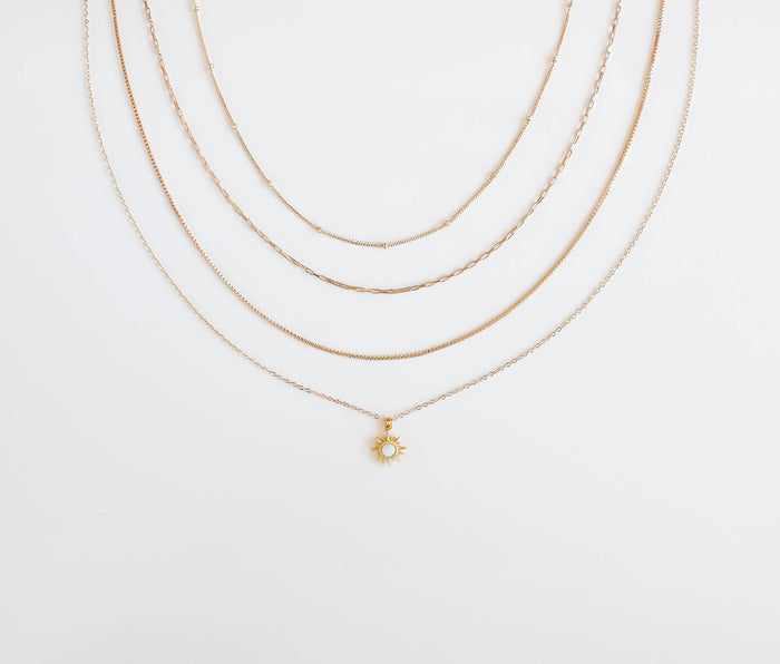 The Cyra Solstice Set by LLUME Jewelry features four delicate gold necklaces on a white background; the shortest displays an opal sun pendant with a central gem, while the others create an elegant layered look.
