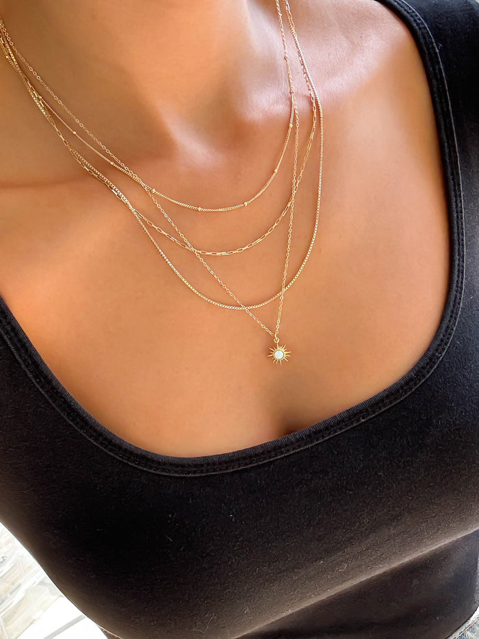 A close-up of a person wearing a black square-neck top and LLUME Jewelry’s Cyra Solstice Set—a gold layered necklace with an opal sun pendant featuring a center stone.