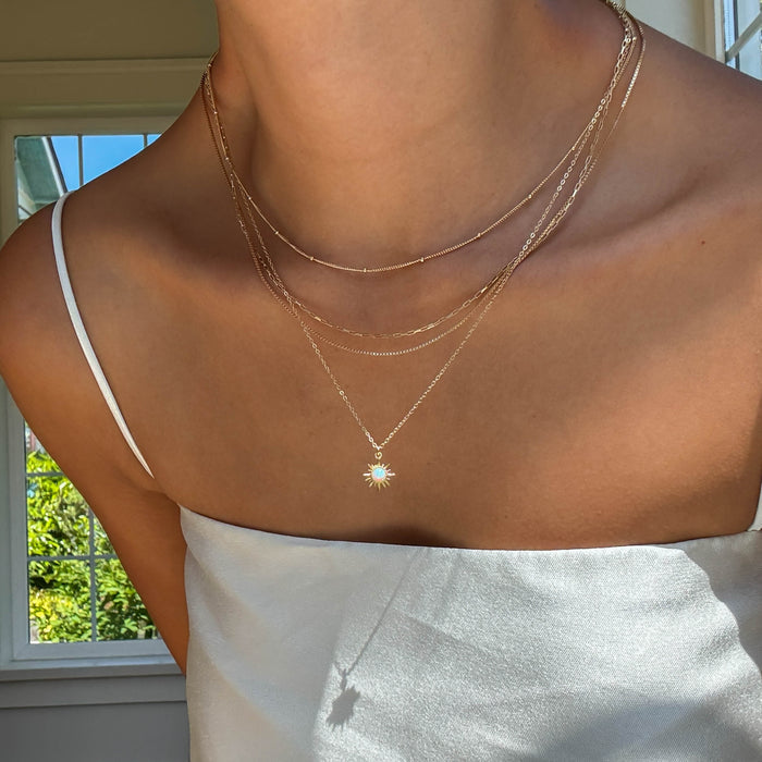 A person stands near a window with greenery outside, wearing a white spaghetti strap top and the LLUME Jewelry Cyra Solstice Set layered necklace.