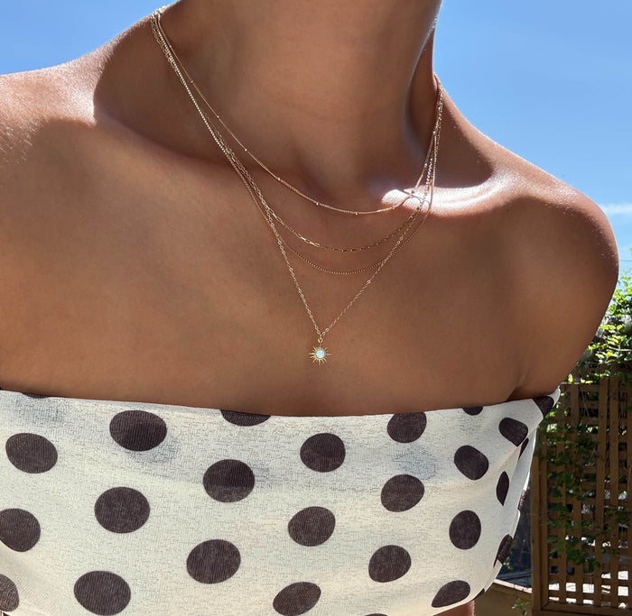 Wearing a white, strapless dress with large black polka dots, a person stands outdoors under a clear blue sky, accessorized with the Cyra Solstice Set by LLUME Jewelry—a gold layered necklace featuring an opal sun pendant.