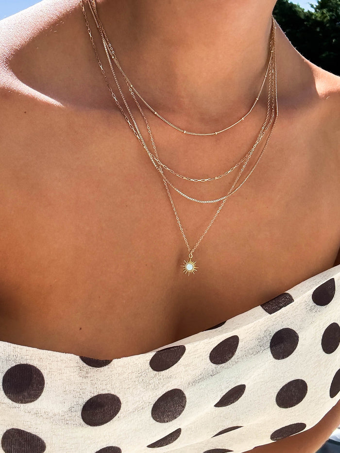 A person outdoors in sunlight wears a white off-shoulder dress with large black polka dots, accessorized with LLUME Jewelry's Cyra Solstice Set featuring a gold layered necklace and opal sun pendant.