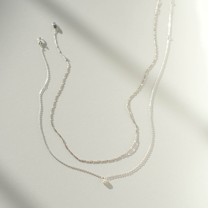 Two delicate pieces of LLUME Jewelry’s Daily Set Silver rest on a smooth surface, casting soft shadows—one a necklace with an elegant charm, the other a simple chain.