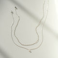Two delicate pieces of LLUME Jewelry’s Daily Set Silver rest on a smooth surface, casting soft shadows—one a necklace with an elegant charm, the other a simple chain.