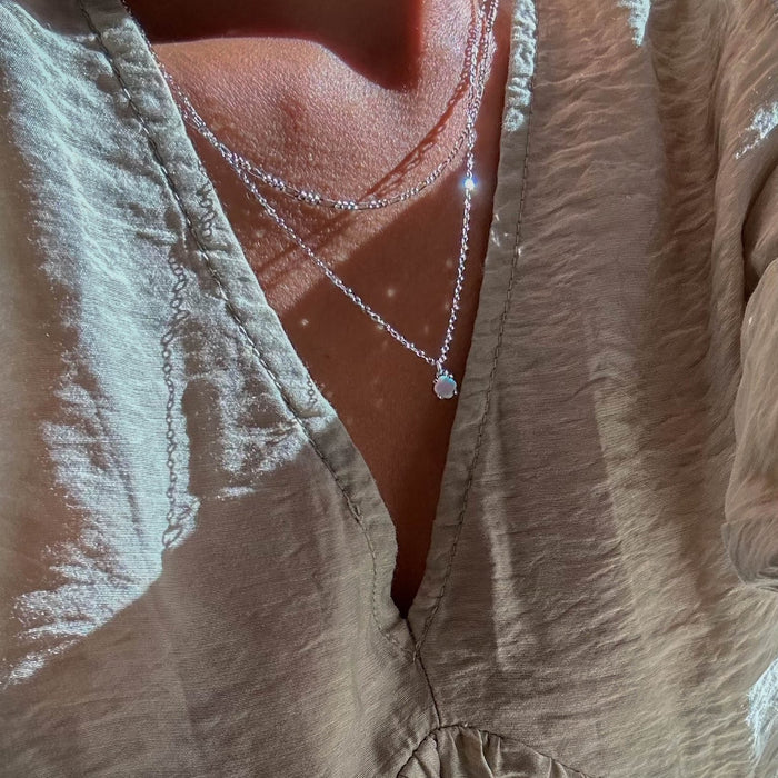 Close-up of a person in a light, textured V-neck blouse, wearing LLUME Jewelry’s Daily Set Silver—two delicate silver necklaces—gleaming in the warm, sunlit scene.