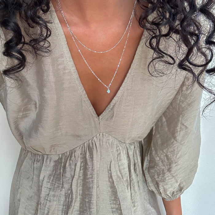 A woman with curly hair wears a light taupe, textured V-neck dress and layered silver jewelry from LLUME Jewelry, including the Daily Set Silver. The cropped image shows her upper body with part of her face out of frame.