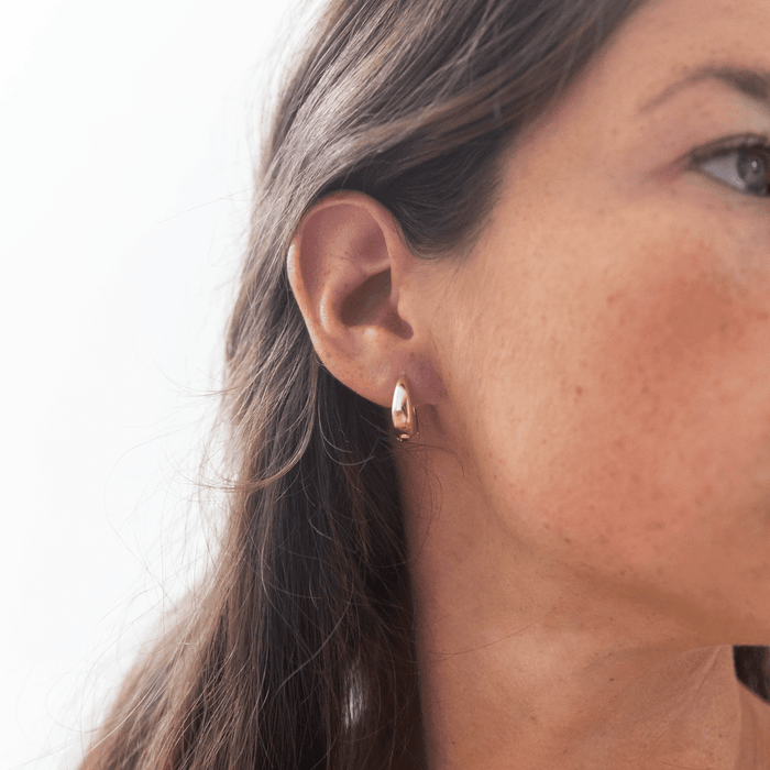 Close-up of a woman wearing LLUME Jewelry's Day-to-Day Hoop, an 18k gold vintage-inspired earring, with straight brown hair. The background is bright and blurred.