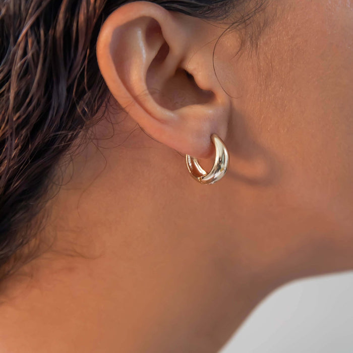 A close-up shows a person with wet, dark hair and light brown skin wearing LLUME Jewelry’s Day-to-Day Hoop earrings. The chunky, shiny 18k gold hoops add a vintage touch, standing out against the softly blurred background.