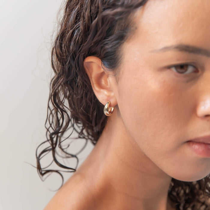A close-up of a woman with curly brown hair wearing the LLUME Jewelry Day-to-Day Hoop—small, shiny 18k gold earrings—against a plain, neutral background.
