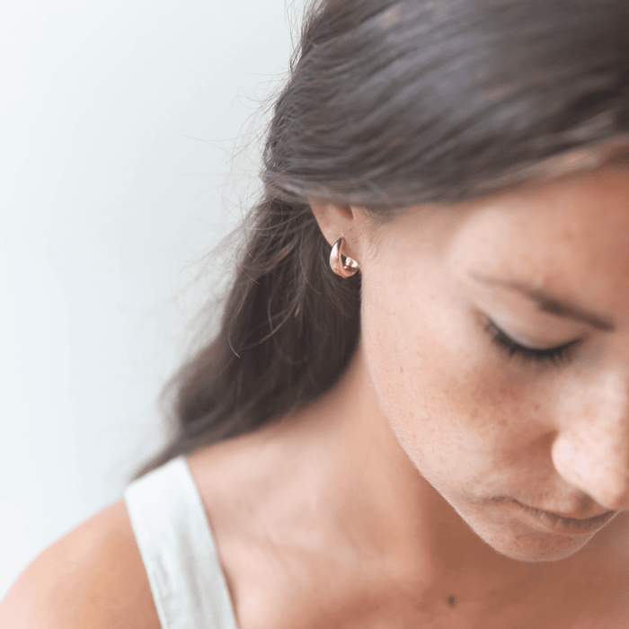 A close-up of a woman with long brown hair wearing the Day-to-Day Hoop by LLUME Jewelry, a pair of delicate 18k gold earrings. She looks down thoughtfully in a softly blurred background, dressed in a sleeveless top.