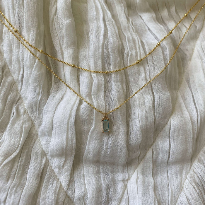 The Daylight Choker Set by LLUME Jewelry features a light blue rectangular pendant necklace paired with a simple gold beaded chain, elegantly layered on textured white fabric.