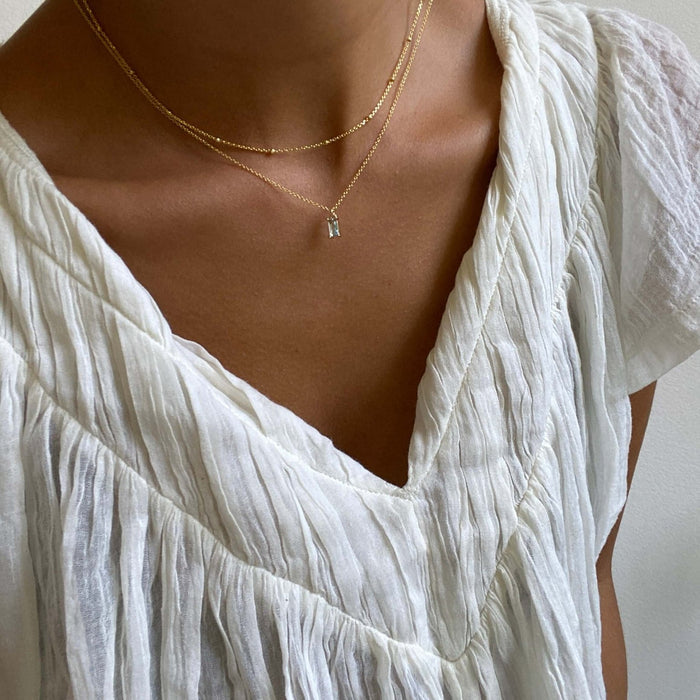 A person wearing LLUME Jewelry’s Daylight Choker Set—featuring a gold beaded chain and delicate layered necklaces with a small pendant—pairs it with a textured white V-neck blouse against a neutral background.