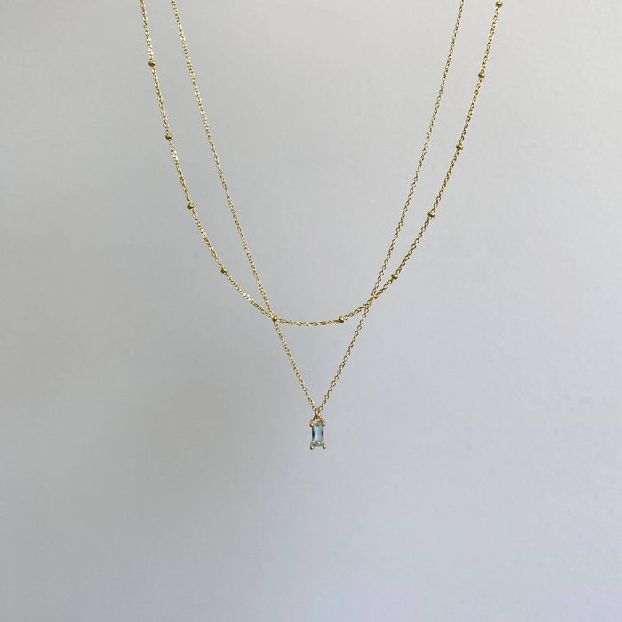The Daylight Choker Set by LLUME Jewelry features fine gold beaded chains—a plain short layer and a longer one with a small rectangular light blue pendant—shown against a simple light background.