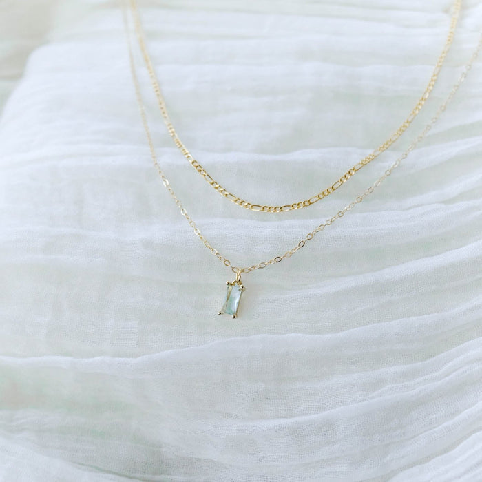 The Daylight Daily Set by LLUME Jewelry features two delicate gold layered necklaces—one with a small light blue stone pendant, the other a simple chain—displayed on soft white fabric and perfect as a jewelry gift.