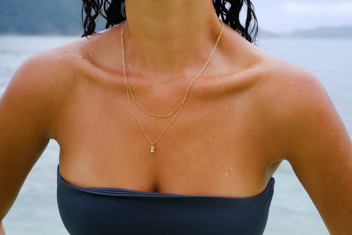 A woman stands in shallow ocean water, droplets on her skin and the blurred shoreline behind her, wearing LLUME Jewelry's Daylight Daily Set layered over a strapless navy blue swimsuit.