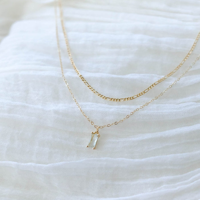 Two delicate gold necklaces from LLUME Jewelry’s Daylight Daily Set rest on white textured fabric; this elegant set features a thin chain and a small rectangular pendant with a clear stone—perfect as a gift or for effortless layering.