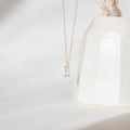 The Daylight Necklace by LLUME Jewelry, featuring a delicate gold-plated chain and a small rectangular clear gemstone pendant, is displayed in front of a white ceramic vase against a soft, minimal background.