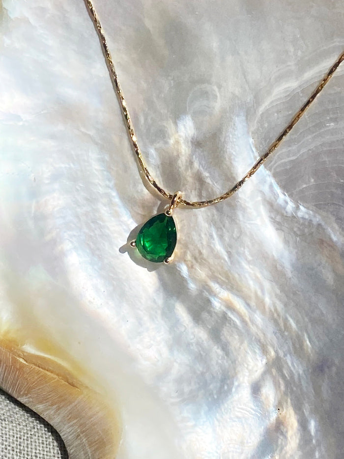 The Delia Necklace by LLUME Jewelry features a teardrop emerald green gemstone pendant on an 18k gold filled chain, displayed on an iridescent shell surface for a soft, elegant look.
