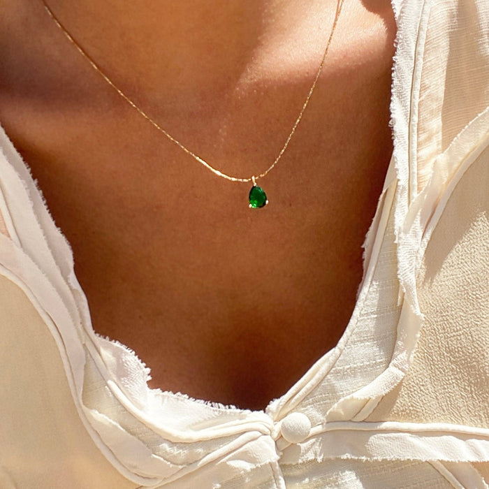 A close-up of a person wearing LLUME Jewelry's Delia Necklace, an 18k gold filled piece with a small oval emerald green pendant, paired with a cream textured top tied at the front.