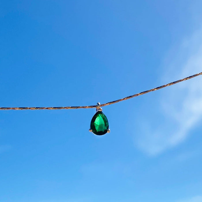 The Delia Necklace by LLUME Jewelry features an emerald green teardrop pendant on a slim 18k gold-filled chain, shown against a clear blue sky.