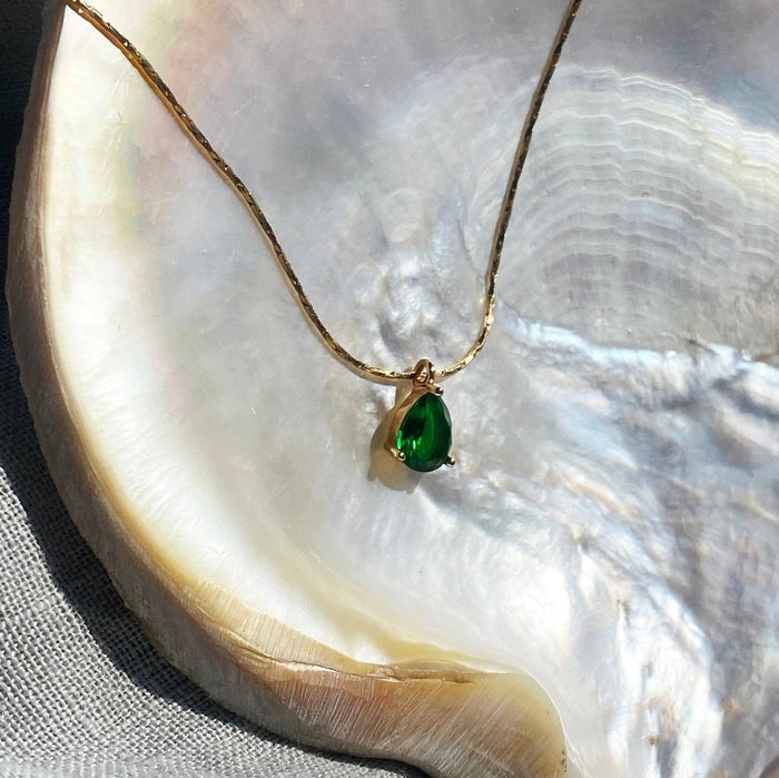 The Delia Necklace by LLUME Jewelry features an emerald green teardrop pendant set in 18k gold filled metal, displayed on a shiny iridescent shell with a textured surface.