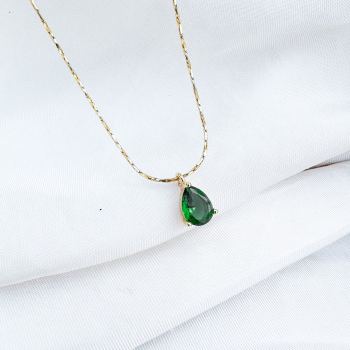 The Delia Necklace by LLUME Jewelry features a teardrop emerald green gemstone pendant on an 18k gold filled chain, elegantly displayed on a white fabric background.