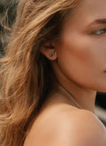 Close-up of a woman with wavy light brown hair wearing LLUME Jewelry’s Destiny Earrings—emerald green studs—and a thin necklace, set against a blurred background.