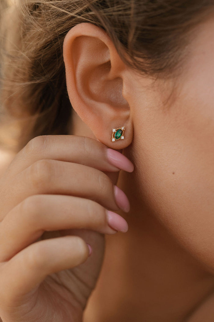 A close-up shows a person wearing LLUME Jewelry’s Destiny Earrings—small gold studs with emerald green gemstones encircled by clear stones—while their pink-manicured hand gently touches their ear.