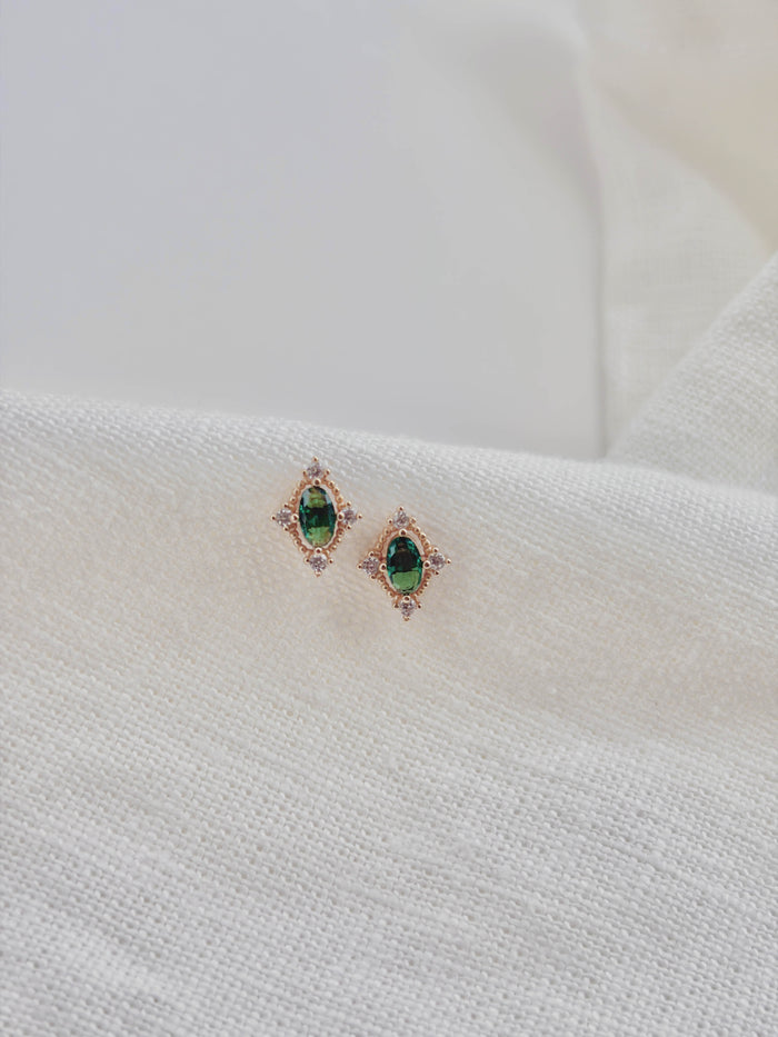 Destiny Earrings by LLUME Jewelry feature gold settings with emerald green studs and clear stones, displayed on a textured white fabric background.