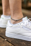 A close-up image of a man's ankle wearing a gray cord anklet with a silver-tone dumbbell charm, paired with white sneakers.