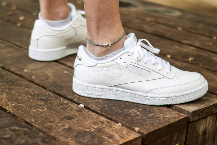 A close-up image of a man's ankle wearing a gray cord anklet with a silver-tone dumbbell charm, paired with white sneakers.