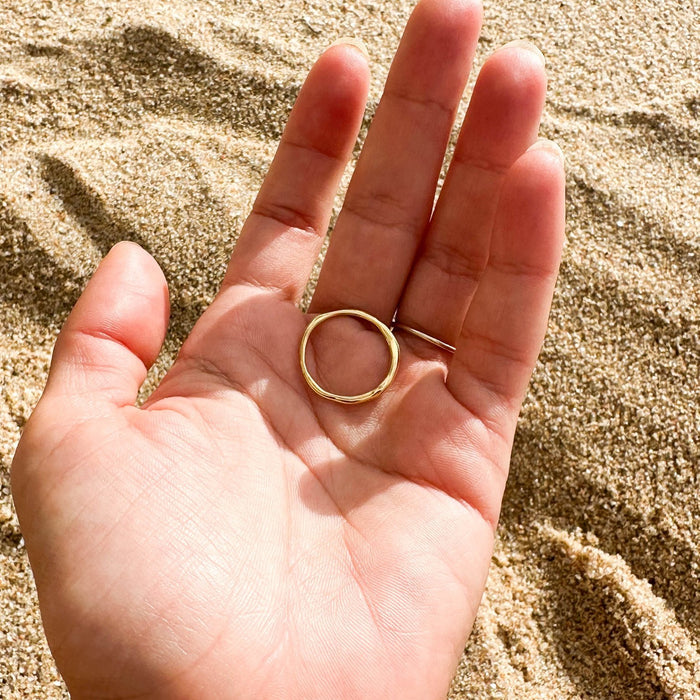 A hand holds the Dune Ring by LLUME Jewelry, a minimalist 14k gold-filled piece ideal for everyday wear, with a sandy beach in the background.
