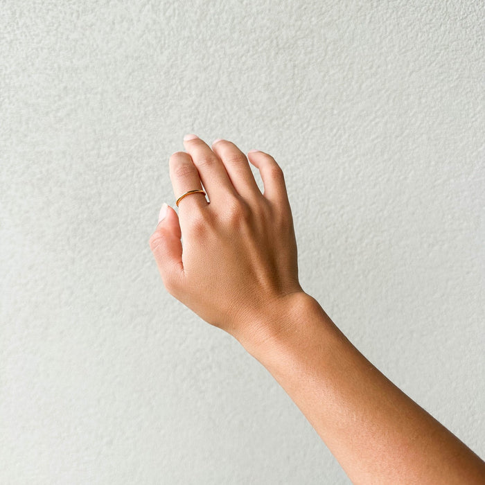 A hand with a light tan complexion is shown against a textured white background, wearing the LLUME Jewelry Dune Ring—a simple 14k gold-filled piece on the middle finger, perfect for daily wear. The relaxed fingers are slightly spread.