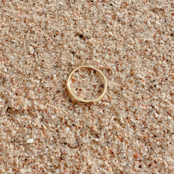 The Dune Ring by LLUME Jewelry, crafted in 14k gold fill, rests on coarse light sand with tiny pebbles—ideal for daily wear or stacking with other rings.