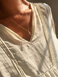 A person wearing the Ella Cross Necklace by LLUME Jewelry—a delicate, gold-plated necklace with a small cross pendant—paired with a cream-colored, lace-trimmed blouse, photographed from the shoulders to upper chest in warm sunlight.