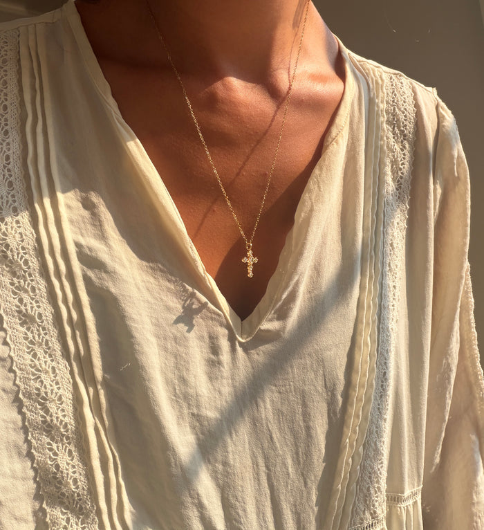 A person wearing a cream-colored, textured blouse and LLUME Jewelry’s Ella Cross Necklace—gold plated and waterproof—stands with sunlight illuminating their skin and clothing, showcasing both style and durability.
